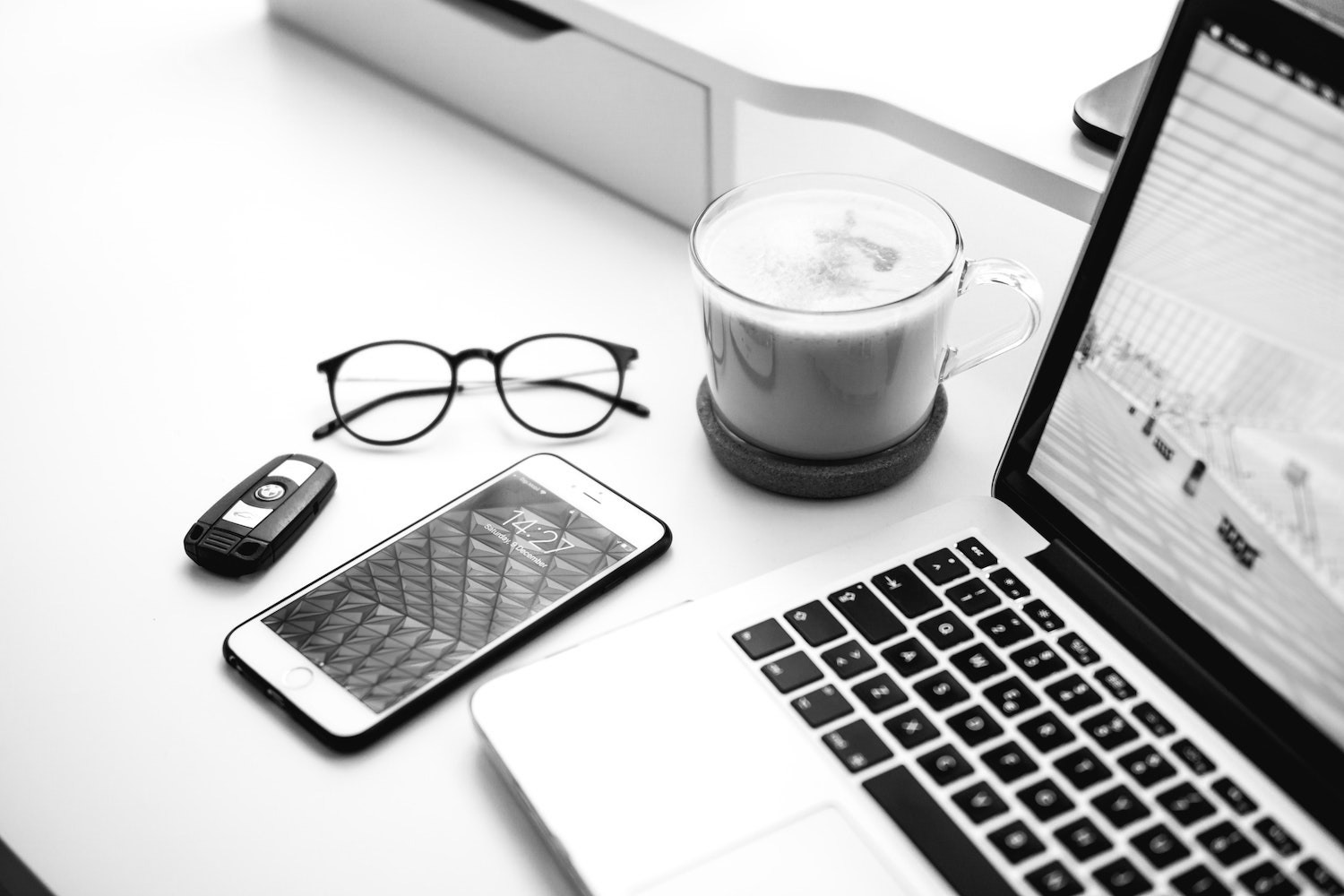 A laptop on a desk next to a cup of frothy coffee, a pair of glasses, a mobile phone and a key fob